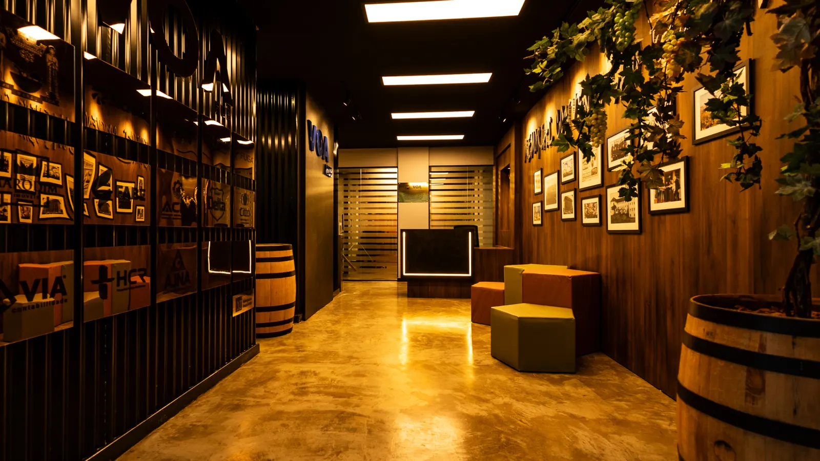 Corredor do Voa Hub com placas de parceiros, quadros, plantas, barris de madeira e bancos coloridos